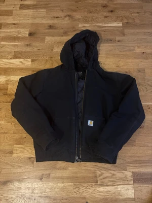 Svart Carhartt hoodiejacka loose fit - Svart hoodiejacka från Carhartt, loose fit i storlek S. Jackan har dragkedja, huva och ribbade muddar. Yttertyget är i 100% bomull och fodret är quiltat för extra värme. Klassisk Carhartt-logga på fickan framtill. Pris kan diskuteras 