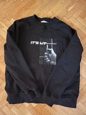 Svart sweatshirt från NA-KD med print - Svart sweatshirt från NA-KD i storlek L med tryck på bröstet som visar en hand med tändare och texten 'IT'S LIT' i 3D-effekt. Tröjan har rund hals och långa ärmar med ribbade muddar. Perfekt för dig som gillar streetstyle och coola prints. Står storlek L men den sitter som M/S
