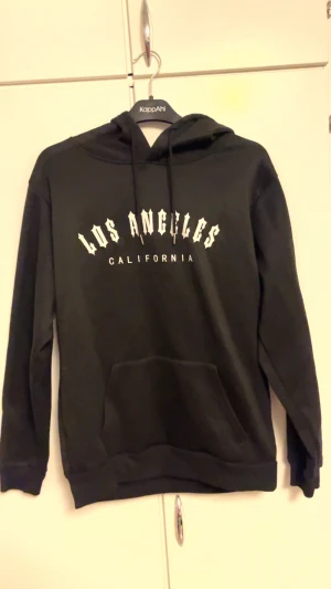 Svart hoodie Los Angeles California - Svart hoodie med trycket 'Los Angeles California' i vitt framtill. Tröjan har huva med snörning och en stor magficka. Långärmad och med ribbade muddar vid ärmslut och nederkant. Perfekt för dig som gillar streetstyle och bekväma plagg.