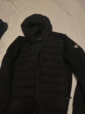 Svart pufferjacka från 1896 Italia - Svart pufferjacka med huva från 1969 Italia. Jackan har quiltad framsida, slät baksida och ärmar, samt en snygg logotyp-patch på vänster ärm. Dragkedja framtill och två sidofickor. Perfekt för dig som gillar stilren och sportig look.