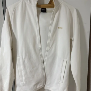 Vit zipjacka från Hugo Boss - Vit långärmad jacka från Hugo Boss med dragkedja framtill och diskret logga på bröstet. Snygga Boss-detaljer längs ena ärmen. Jackan har två fickor och är tillverkad i mjuk bomullsmix för en clean och fräsch look. Storlek M