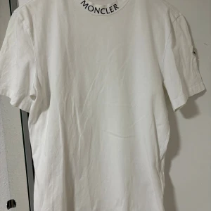 Vit Moncler t-shirt med logga - Snygg vit t-shirt från Moncler i bomull, med svart Moncler-tryck runt halsen och broderad logga på ärmen. Klassisk passform och korta ärmar. Storlek Xs men passar S och M