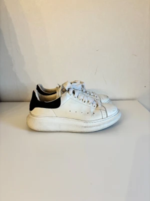 Alexander McQueen vita sneakers - Säljer ett par vita sneakers från Alexander McQueen med svart mockadetalj på hälen. Skorna har bred platt sula, rund tå och klassisk snörning. Skorna är väl använda och defekter syns på bilderna. Har dessvärre inget äkthetsbevis.
