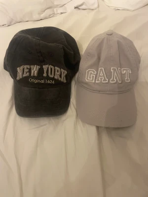 Svart New York keps & beige GANT keps - Två snygga kepsar: en svart Robin Ruth med broderad 'NEW YORK Original 1624' framtill och en beige GANT med vit broderad logga. Båda har klassisk böjd skärm och justerbar passform. GANT-kepsen har rutigt band på insidan för extra stil.