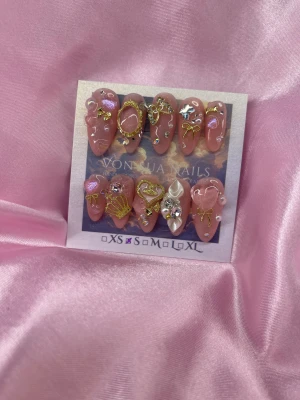 CoCo (S);Medium Almond - Set med handgjorda lösnaglar i rosa med glansig finish, dekorerade med 3D-accessoarer som pärlor, strass, hjärtan, rosetter och guldfärgade detaljer. Naglarna är en medium mandelform och finns i S: 15 mm, 11,5 mm, 12 mm, 11 mm, 9 mm. Perfekt för dig som vill ha extra glam och unik design.