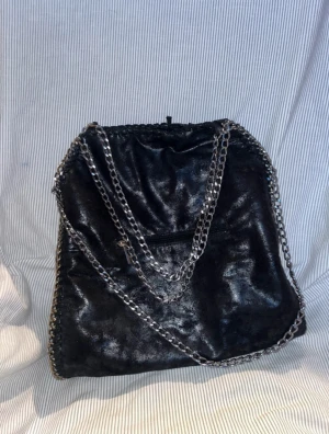 Svart axelväska med kedjedetaljer - Trendig svart axelväska i skinnimitation med glansig finish och silverfärgade kedjor som axelrem. Väskan har snörning upptill och dekorativa sömmar längs kanterna. Insidan är fodrad med mönstrat tyg och har en innerficka med dragkedja.