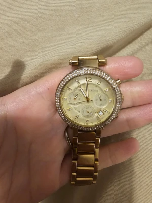 Guldig Michael Kors klocka med stenar - Säljer en snygg guldig klocka från Michael Kors med rund urtavla och glittrande stenar runt ramen. Klockan har länkarmband i metall, flera små urtavlor och datumvisning. Perfekt accessoar för dig som gillar bling och stilren design.