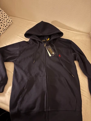Marinblå hoodie från Polo Ralph Lauren - Snygg marinblå hoodie från Polo Ralph Lauren med dragkedja och huva. Tröjan har långa ärmar, snörning vid huvan och en röd broderad logga på bröstet. Tillverkad i mjuk bomull och har praktiska fickor framtill. Perfekt för chill dagar.
