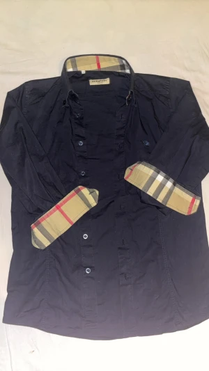 Svart/Mörkblå Burberry skjorta med rutiga detaljer - Svart/Mörkblå skjorta från Burberry med klassiska rutiga detaljer i beige, rött och svart på insidan av kragen och ärmsluten. Skjortan har knappar framtill och broderad logga på bröstet. Snygg och tidlös design i bomull.Det står L men sitter riktigt bra som M elr långa personer med S kan funka.Pris kan diskuteras och byte med. Skriv om ni har några frågor:)