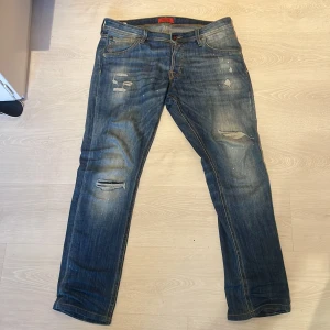 Blå jeans från Slim glenn Jack & Jones - Säljer ett par blå jeans från Jack & Jones med slitna detaljer och små färgstänk. Klassisk femficksmodell med rak passform och snyggt tvättad look. Jeansen har normal midja och är tillverkade i vanligt jeans matrial för en skön känsla.