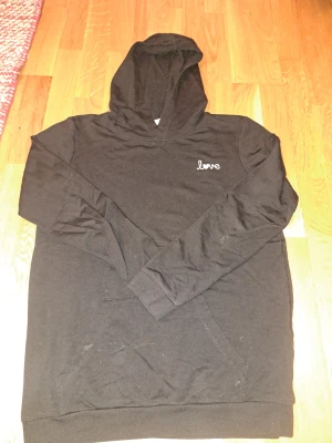 Svart hoodie med love-tryck - Svart hoodie med vit broderad text 'love' och ett hjärta på bröstet. Tröjan har huva, långärmade ärmar och en klassisk magficka framtill. Materialet känns som mjuk bomull och passformen är normal. Perfekt för chill dagar och enkel att matcha med jeans eller joggers.