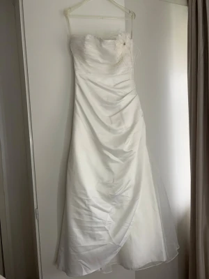 Vit axelbandslös bröllopsklänning  - Säljer en elegant vit axelbandslös långklänning i satin med draperad framsida och avtagbar blomdetalj med fjädrar och pärlor. Medföljer en matchande liten väska med rosett i samma material. Finns några skavanker som finns på bilderna, går förmodligen att tvätta bort. Priset kan diskuteras!