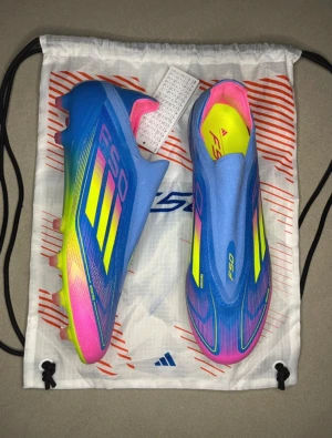 Adidas F50 Elite Laceless AG - Säljer ett par Adidas F50 fotbollsskor med en riktigt fet färgkombo i blått, neon-gult och rosa. Skorna har en strömlinjeformad design med platt sula och rund tå, samt mönstrade detaljer och Adidas-logga på sidan. Ovandelen är i syntetmaterial för lätt känsla på planen.