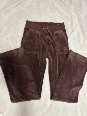 Bruna mjukisbyxor Juicy Couture - Snygga bruna mjukisbyxor från Juicy Couture med broderad JC-logga på fickan. Byxorna har snörning i midjan och är gjorda i mjuk sammet som känns lyxig mot huden. Perfekta för chill dagar och har en klassisk, relaxed passform.