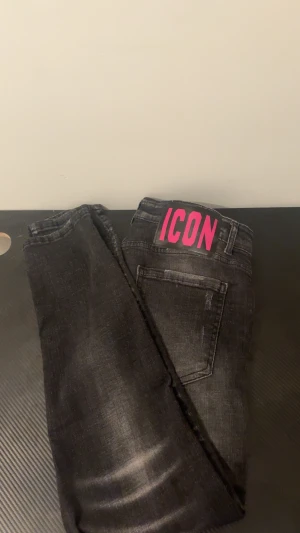 Svarta Dsquared2 ICON jeans, storlek 52 - Säljer ett par svarta jeans från Dsquared2 med coola slitningar och blekta detaljer. På bakfickan finns en stor rosa ICON-logga som sticker ut. Jeansen har klassisk femficksdesign och är gjorda i jeansmaterial med en slim passform.