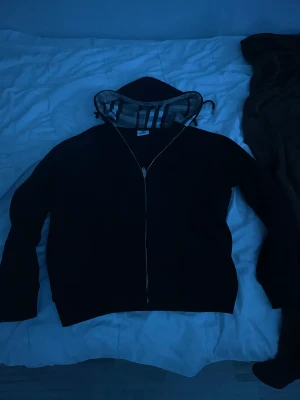 Svart hoodie från Burberry med dragkedja - Svart hoodie från Burberry med klassisk rutmönstrad insida på huvan och hel dragkedja framtill. Tröjan har långa ärmar och en normal passform. Perfekt för dig som vill ha en stilren och lyxig look med ikoniska Burberry-detaljer.