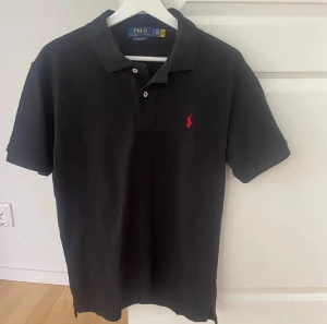 Svart pikétröja från Polo Ralph Lauren - Svart pikétröja från Polo Ralph Lauren med klassisk krage och två knappar framtill. Tröjan har korta ärmar och den ikoniska röda Polo-loggan broderad på bröstet. Tillverkad i mjuk bomull som är skön mot huden och passar perfekt till jeans eller shorts.