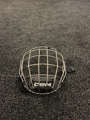 CCM hockeygaller i silver - Säljer ett robust hockeygaller från CCM i silverfärgad metall med svart hakskydd och justerbara remmar. Gallret har en klassisk fyrkantig form och passar perfekt till hockeyhjälm för extra skydd på isen.