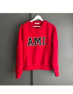 Röd sweatshirt från AMI  - Säljer en röd sweatshirt från AMI med stort svart och vitt tryck på bröstet. Tröjan har rund hals, långa ärmar och ribbade muddar vid ärmslut och nederkant. Perfekt för dig som gillar streetwear och vill sticka ut med en snygg logga.