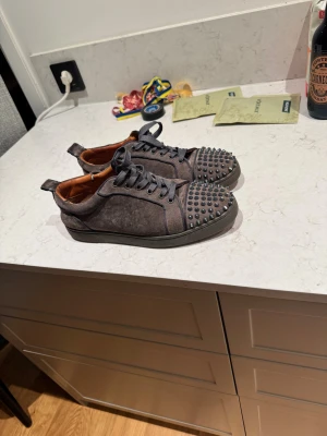 Grå mocka loubitin med nitar - Unika grå sneakers i mocka med coola svarta nitar på tån och klassisk snörning. Insidan är brun och yttersulan har en fet röd färg som sticker ut. Snygg rund tå och platt sula, perfekt för dig som vill ha något som syns och känns lyxigt.