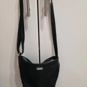 Svart axelväska från Don Donna - Snygg svart axelväska från Don Donna med stilren design och silverfärgad logga framtill. Väskan har flera praktiska fack med dragkedja och är tillverkad i mjukt syntetmaterial med polyesterfoder. Perfekt storlek för att få plats med allt du behöver.