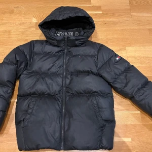 Svart dunjacka från Tommy Hilfiger, strl 164 - Snygg svart vinterrjacka strl 164 från Tommy Hilfiger med huva. Varm dunfyllning perfekt för kyliga dagar. Rätt stor i storleken o fint skick.