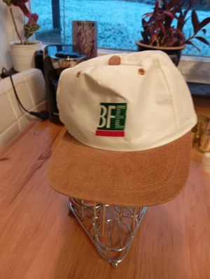 Ny BFE keps med manchester - Snygg keps från BFE med vit krona och brun skärm i manchester. Framsidan har en broderad BFE-logga i grönt, vitt och rött. Perfekt för dig som gillar retrostil och vill sticka ut med en unik accessoar.