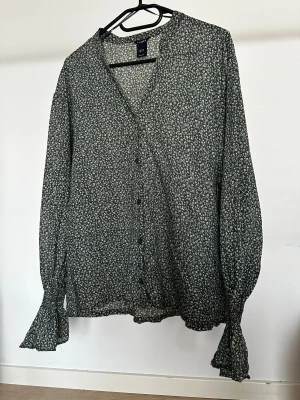 Blus från Lindex - Blommig blus från Lindex i grönt med vita små blommor. V-ringning, knappar framtill och långa ärmar med volang vid ärmslutet. 