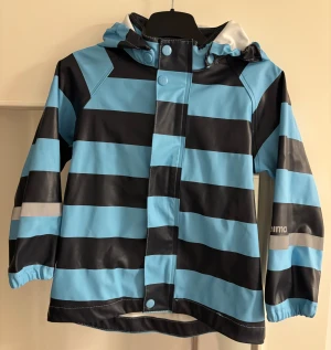 Randig regnjacka från Reima blå/svart - Snygg regnjacka från Reima med breda blå och svarta ränder, reflexdetaljer och avtagbar huva. Jackan har tryckknappar framtill och är tillverkad i vattentätt syntetmaterial. Perfekt för blöta dagar och håller dig torr med stil.