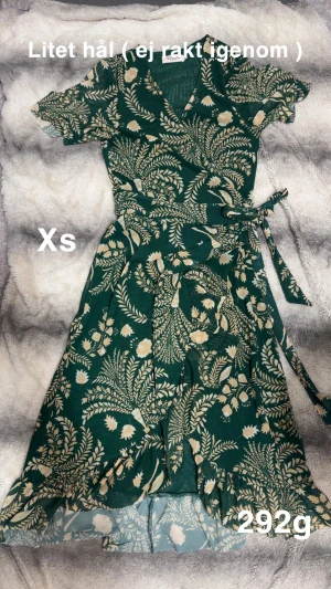 Grön blommig omlottklänning Soaked - Supersöt grön omlottklänning från Soaked in Luxury med beige blommönster. Klänningen har volangdetaljer nertill, korta ärmar och knytband i midjan. Perfekt för dig som gillar en feminin och flowy stil.🌸 Litet hål i inre delen av tyget. Storlek xs