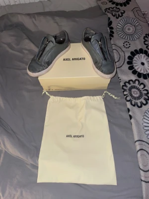 Grå sneakers Axel Arigato med dragkedja - Snygga grå sneakers från Axel Arigato i skinn med cool dragkedjedetalj framtill istället för klassisk snörning. Skorna har vit platt sula och en rund tå. Medföljer originalkartong och dustbag. Perfekt för dig som gillar minimalistisk och modern stil.