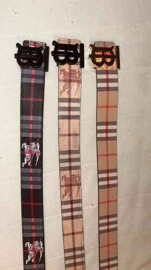  Burberry bälte - Snyggt bälte med klassiskt rutigt Burberry-mönster i beige, brunt och rött. Bältet har en stor metallspänne med B-logga i guld eller svart. Materialet är skinnimitation med tryckt mönster och insida i svart. Har extra B guld stämpel.