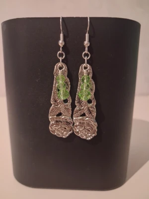 Egentillverkade örhängen återbruk  - Unika hängande örhängen skapade av 2st skedar i rostfritt stål format som en ros. Örhänget pryds av gröna pärlor. Öronkroken är nickelsäker och aldrig använda. Blir en elegant och unik accessoar 