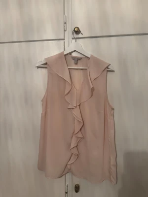 Ljusrosa blus med volang från H&M - Säljer en ljusrosa blus från H&M med snygg volang längs fram och bred krage. Blusen är ärmlös och har en flowy passform som ger en chill och feminin vibe. Perfekt för dig som gillar romantiska detaljer och mjuka färger.
