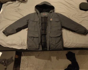 Grå parkajacka från Fjällräven - Säljer en skit snygg Fjällräven jacka med huva och flera stora fickor framtill samt skit bra innerfickor. Jackan har dragkedja och knappar, samt klassisk Fjällräven-logga på bröstet. Perfekt för kalla dagar.  Ny pris runt ca 5-6 tusen.   Säljer pga ny vinter jackla.     Ba skriva om ni undrar nått.   