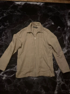 Beige overshirt från J.Lindeberg - Snygg beige overshirt från J.Lindeberg i storlek M. Jackan har klassisk krage, hel dragkedja framtill och är tillverkad i ett lätt linnematerial. Perfekt för dig som gillar stilrena och enkla plagg med clean look.
