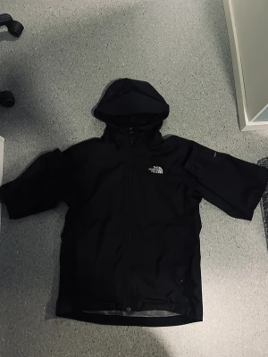 Svart vindjacka från The North Face - Svart vindjacka från The North Face med huva och dragkedja framtill. Jackan har elastiska muddar vid ärmslut och logga på både bröstet och ryggen. Perfekt för blöta dagar och har en clean, sportig look. Materialet är syntet och jackan är långärmad.