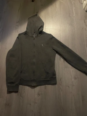 Mörkgrå hoodie från Polo Ralph Lauren - Mörkgrå hoodie från Polo Ralph Lauren med dragkedja framtill och klassisk broderad logga på bröstet. Tröjan har huva, känguruficka och långa ärmar. Perfekt för chill dagar och enkel att matcha med jeans eller joggers. Hör av dig vid fundering 🤔. Pris kan sänkas vid snabb affär