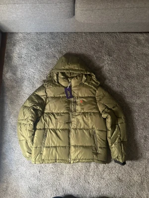 Olivgrön pufferjacka från Polo Ralph Lauren - Snygg olivgrön pufferjacka från Polo Ralph Lauren med huva och två dragkedjeförsedda fickor. Jackan har broderad röd logga på bröstet och grov dragkedja framtill. Materialet är glansigt syntet och jackan är quiltad för extra värme.