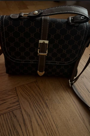 Brun axelväska - Snygg brun axelväska  med klassiskt monogrammönster och guldfärgade detaljer. Väskan har justerbar axelrem, dekorativt spänne framtill och flera fack med dragkedja inuti. Tillverkad i skinn med kontrastsömmar för en lyxig känsla.