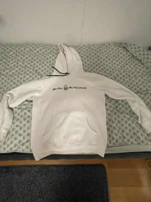 Vit hoodie från Sail Racing - Vit hoodie från Sail Racing med svart logga på bröstet. Klassisk huva med svarta snören och stor magficka framtill. Perfekt för dig som gillar en enkel och stilren look. Mjuk insida och normal passform.