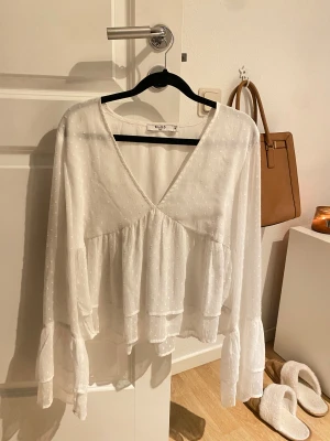 Vit blus med volang från NA-KD - Luftig vit blus från NA-KD med v-ringning och volangdetaljer. Blusen har långärmade ärmar med lätt volym och diskret prickigt mönster i tyget. Perfekt för en fräsch och stilren look.