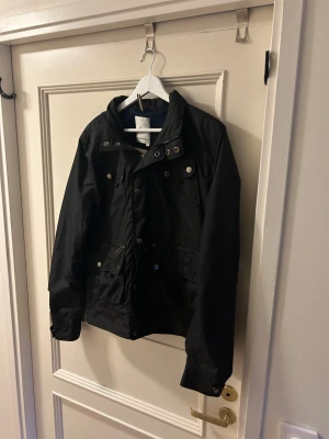 Svart vindjacka från J.Lindberg - Svart vindjacka från J.Lindberg med flera fickor framtill, både med dragkedja och knappar. Det är storlek L men passar mer M.Jackan har ståkrage med tryckknappar och randigt foder på insidan. Bra kvalitet och jackan är långärmad med justerbara muddar. Knappt använd. Skriv om ni undrar något. Priset är inte hugget i sten 