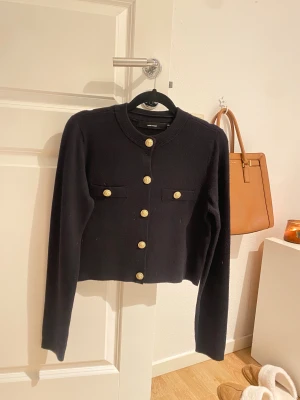 Svart kofta med guldknappar från Vero Moda - Stilren svart kofta från Vero Moda med rund hals och snygga guldknappar framtill. Koftan har två dekorativa fickor på bröstet och är långärmad. Perfekt för dig som gillar klassiska detaljer och vill ha en enkel men elegant look.