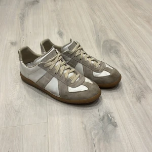 Maison margiela  - Snygga sneakers i beige och vitt med coola paneler i mocka och skinn. Dom har 2 små defekter som ni ser på bild 4 och 5 då det har lossnat lite vit färg men inget man ser eller tänker på vid andväning. Perfekt för dig som gillar stilrena och neutrala färger med en retrovibe. Självklart äkta! Dustbag tillkommer!