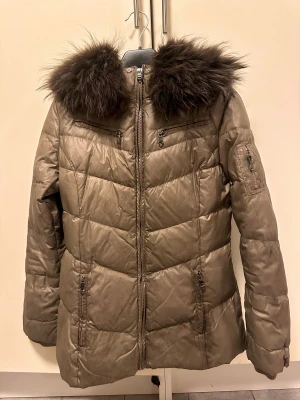 Beige/brun dunjacka med pälsdetalj - Snygg beige/brun pufferjacka med fluffig mörkbrun äkta päls runt huvan. Jackan har flera dragkedjeförsedda fickor och quiltad design som ger en cool look. Perfekt för kalla dagar med sin tjocka och varma känsla. Långärmad och med normal passform stl 34, jackan är fin och hel men färgen är lite ”sliten” på dragkedjorna 
