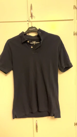 Marinblå pikétröja Hampton Republic - Snygg marinblå pikétröja från Hampton Republic i storlek S. Klassisk krage, korta ärmar och två knappar framtill. Tröjan är gjord i mjuk bomull och har en stilren look som funkar till många olika outfits.