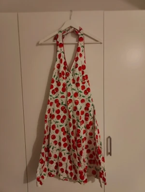 Retro Körsbärsklänning - Vit klänning från Voodoo Vixen med rött körsbärsmönster och svarta prickar. Klänningen har halterneck, v-ringning och en luftig, svängande kjol. Perfekt för dig som gillar retrostil och färgglada prints!