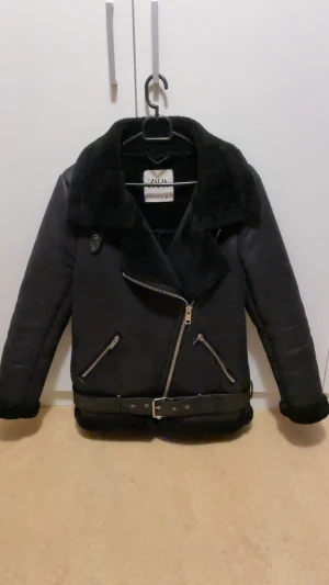 Svart jacka från Zara - Snygg svart jacka från Zara med dragkedjor i silver och bälte. Jackan har mjukt foder och detaljer i päls vid krage och ärmslut. Perfekt för kalla dagar och ger en cool look. Jackan e köpt för 900kr och säljs för 350kr. Storlek XS men passar även S
