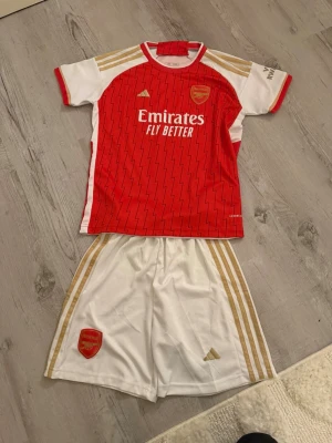 Arsenal matchställ Adidas röd/vit - Säljer ett Arsenal matchställ från Adidas med kortärmad tröja i rött och vitt, guldiga detaljer och zigzag-mönster. Tröjan har klubbmärke, Emirates-tryck och 'Visit Rwanda' på ärmen. Tillhörande vita shorts med guldiga ränder och Arsenal-logga. Materialet är lätt polyester.